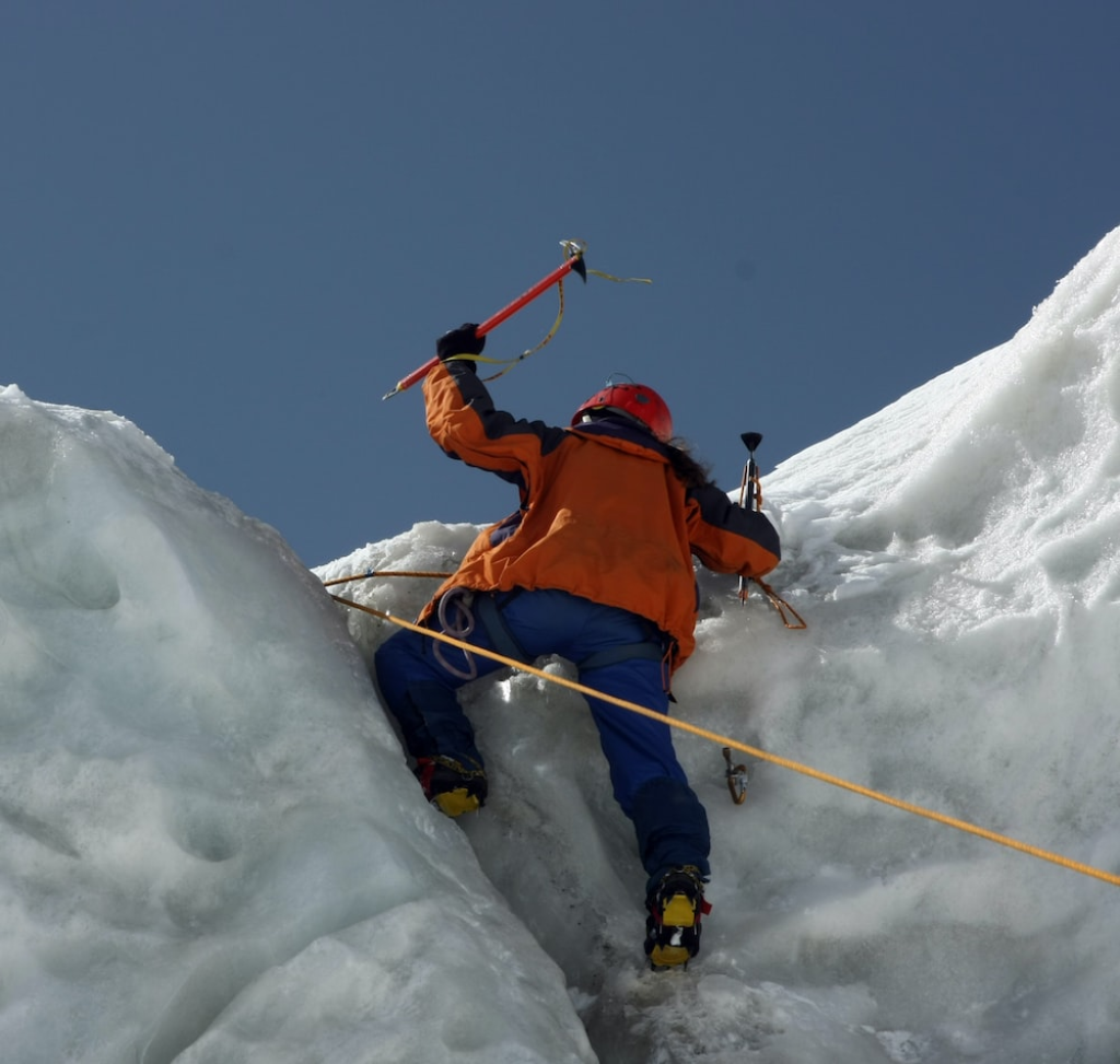An ice climber in an orange jacket using ice axes