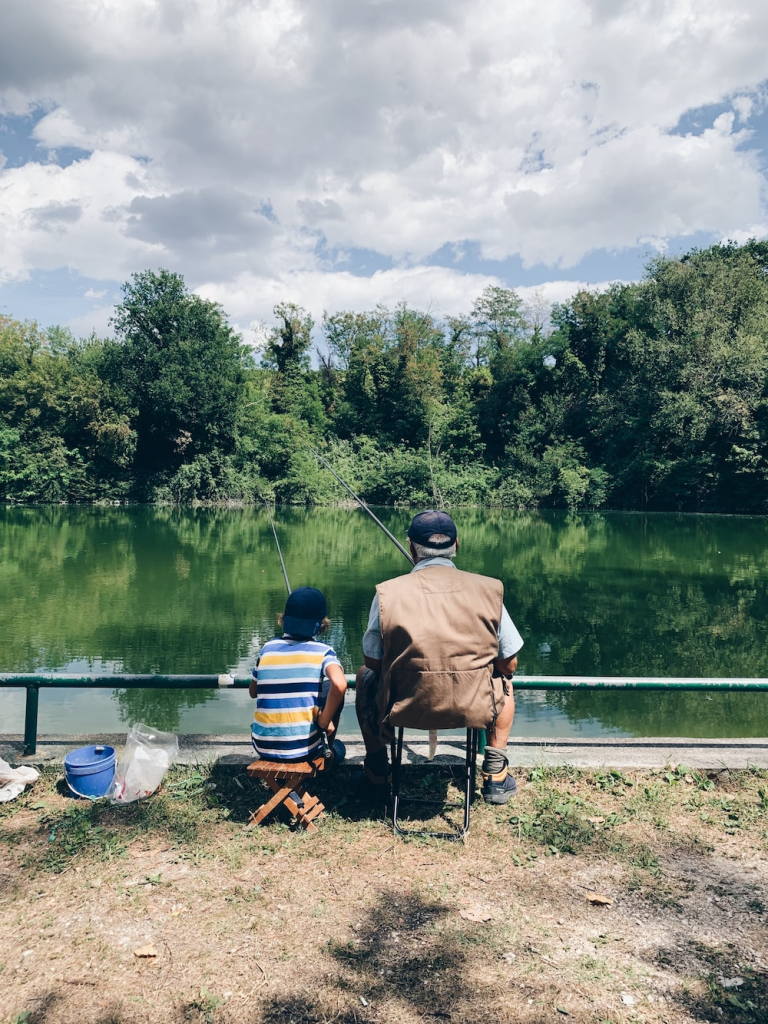 A grand parent and a child sitting together fishing at the edge of a freshwater body.