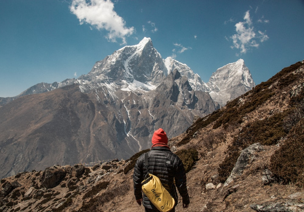 A climber with a yellow backpack and red beanie standing in front of mountains