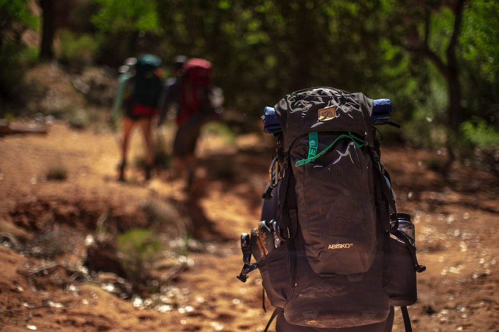 A backpack with people walking behind it on a trail with a sleeping bag.
